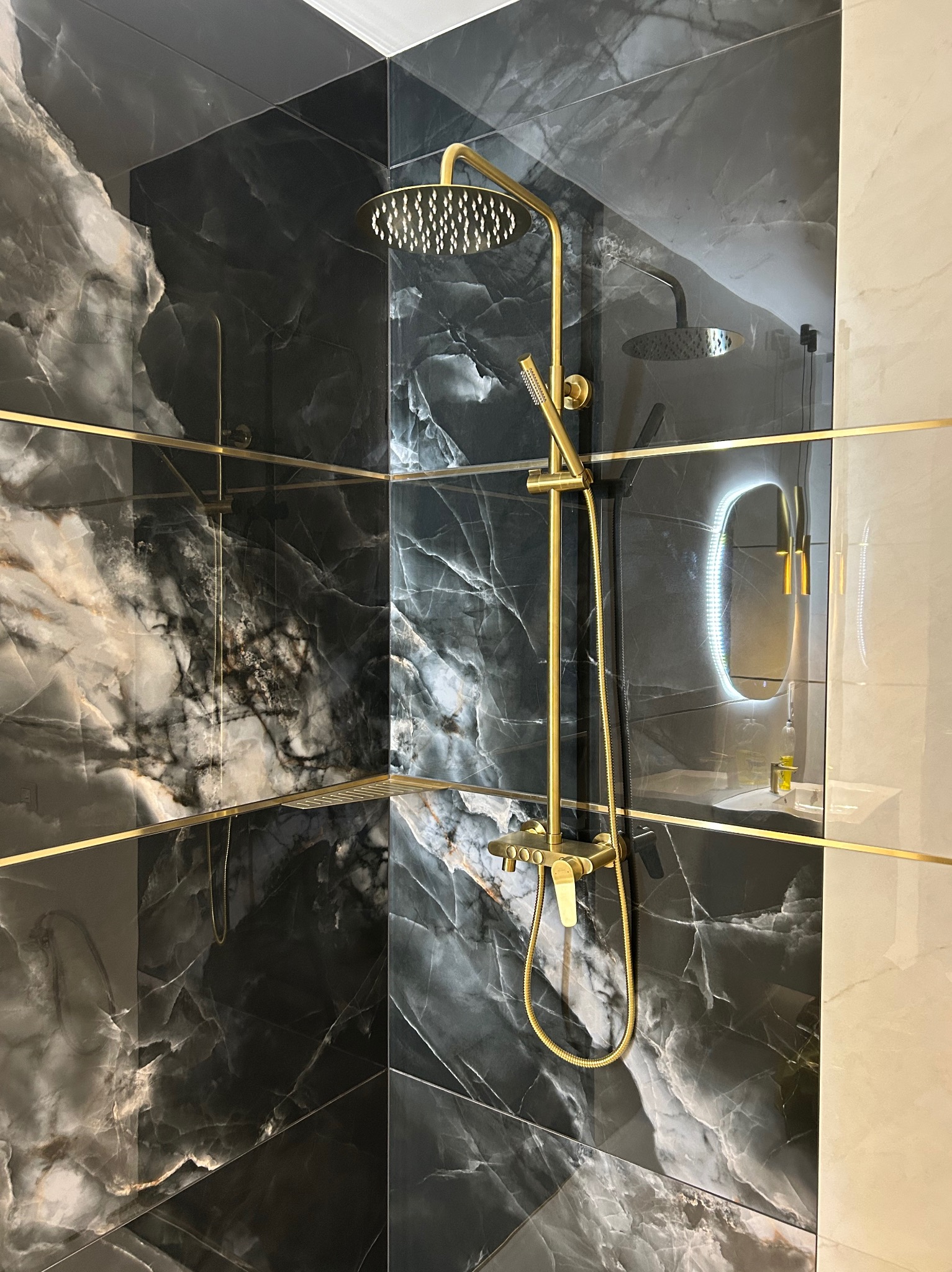 Elegancka kabina prysznicowa z czarnymi płytkami imitującymi marmur, złotą armaturą i podświetlanym lustrem. Nowoczesny design łazienki.