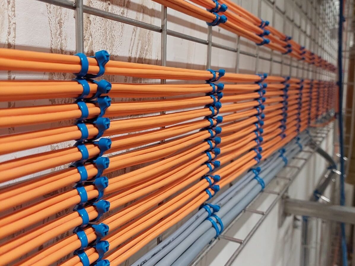 Równolegle ułożone, pomarańczowe i szare przewody elektryczne, przymocowane niebieskimi uchwytami do metalowej korytki kablowej, zamocowanej na ścianie o jasnej strukturze.