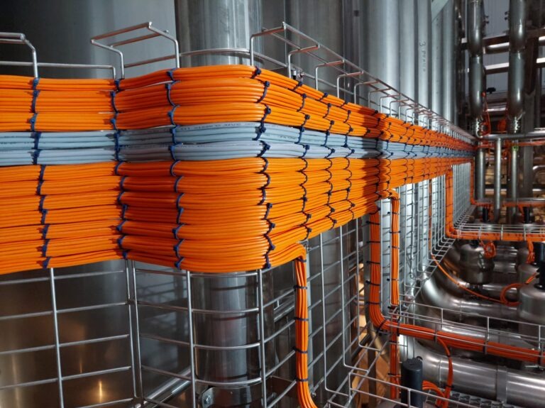 Równolegle ułożone pomarańczowe i szare przewody w metalowej korycie kablowej, zabezpieczone niebieskimi i czarnymi opaskami zaciskowymi, na tle przemysłowych zbiorników ze stali nierdzewnej i rur