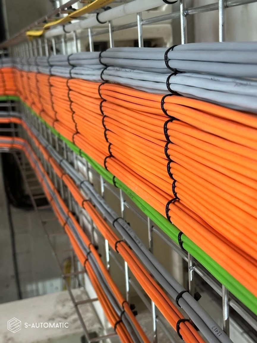 Równolegle ułożone kable elektryczne w korytkach kablowych, przewody w kolorach pomarańczowym, szarym i zielonym, zabezpieczone czarnymi opaskami zaciskowymi.