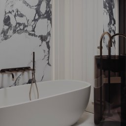 S STUDIO Sandra Kluczek - Nowoczesna łazienka z wolnostojącą białą wanną, baterią w kolorze szczotkowanego złota i umywalką na postumencie z ciemnego szkła. Tło stanowi ściana z imitacją marmuru i biała boazeria.