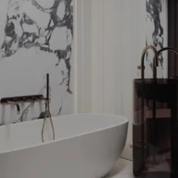 Nowoczesna łazienka z wolnostojącą białą wanną, baterią w kolorze szczotkowanego złota i umywalką na postumencie z ciemnego szkła. Tło stanowi ściana z imitacją marmuru i biała boazeria.