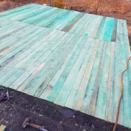 Widok z góry na drewnianą konstrukcję dachu pokrytą zieloną impregnacją, z widocznym młotkiem, narzędziami i liną na czarnym podłożu dachowym.