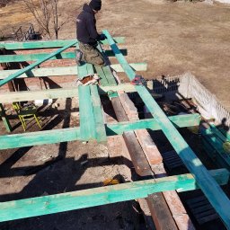 Michał Słomian - Budowa więźby dachowej z pomalowanymi na zielono elementami, widok z góry na dekarza w czapce i kurtce, pracującego na dachu z czerwonej cegły, z zielonym krzesłem i narzędziami w tle.