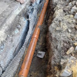 Michał Słomian - Pomarańczowa rura kanalizacyjna ułożona w wykopie ziemnym, zabezpieczonym geowłókniną; widoczne dłonie robotnika i kielnia.