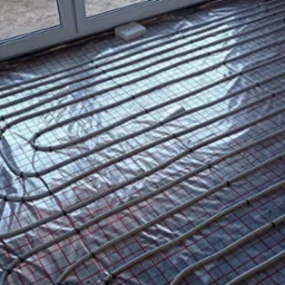 Białe rurki ogrzewania podłogowego ułożone w pętle na srebrnej folii z czerwoną siatką przed wylewką, widok z góry.