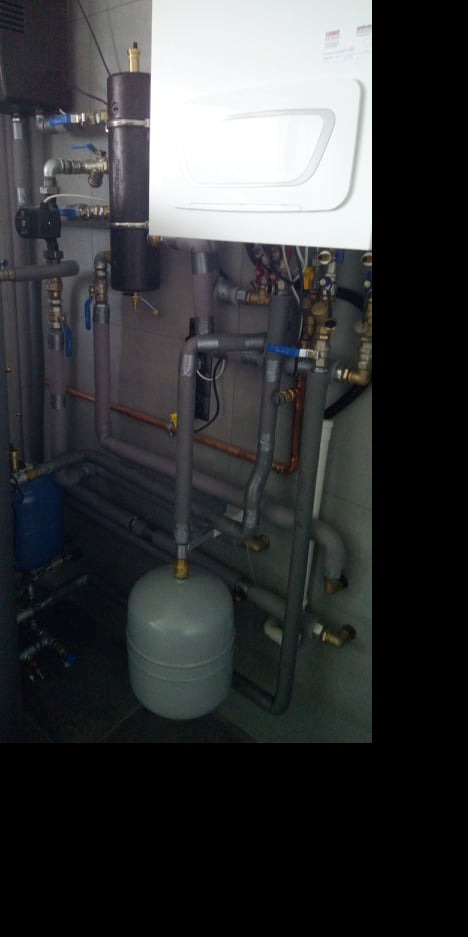 Kompleksowa instalacja grzewcza z białym piecem, czarnym zbiornikiem i szarymi rurami, widoczne liczne zawory i złączki mosiężne.