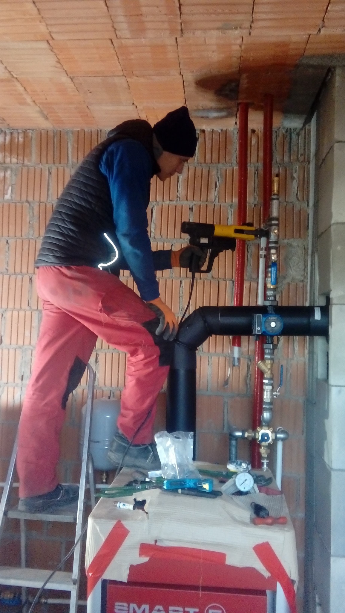 Hydraulik na drabinie montuje rury instalacji centralnego ogrzewania przy użyciu specjalistycznego narzędzia do zgrzewania, widoczne elementy instalacji, manometr i narzędzia na prowizorycznym...