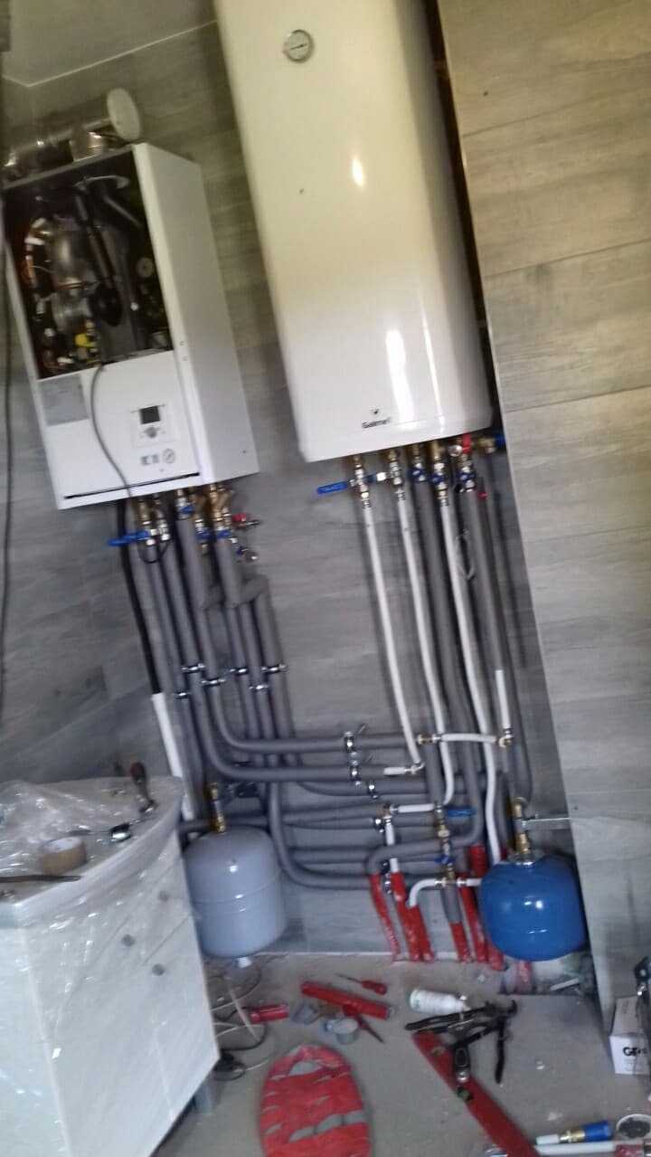 Dwa białe bojlery wiszące na ścianie z szarych płytek imitujących drewno, liczne rury w izolacji, małe zbiorniki wyrównawcze, narzędzia hydrauliczne i akcesoria na podłodze, widok z dołu.