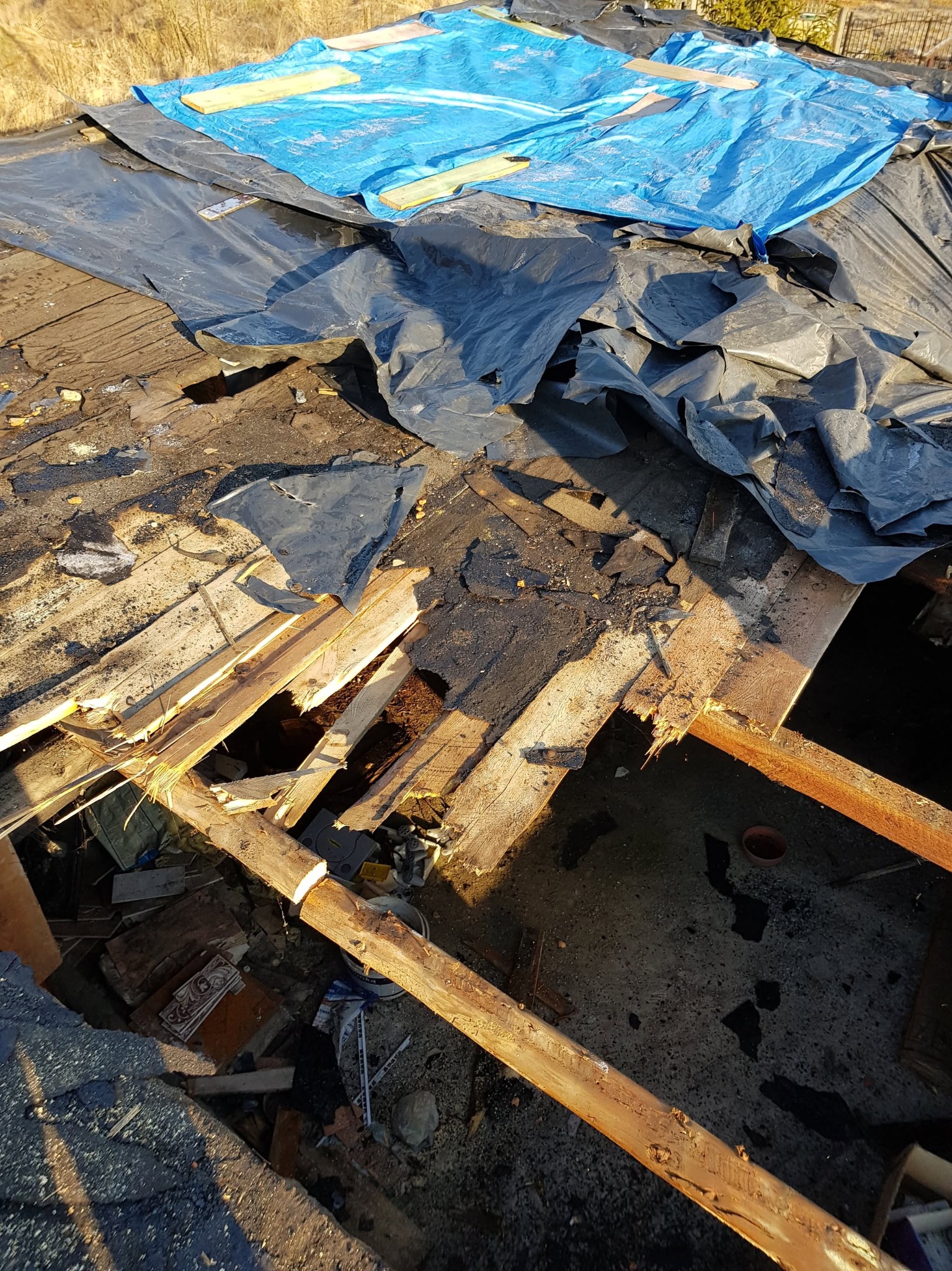 Zdjęcie uszkodzonego, częściowo odkrytego dachu z widocznymi elementami konstrukcyjnymi, zabezpieczonego niebieską i czarną plandeką, z gruzem i odpadami widocznymi wewnątrz budynku.