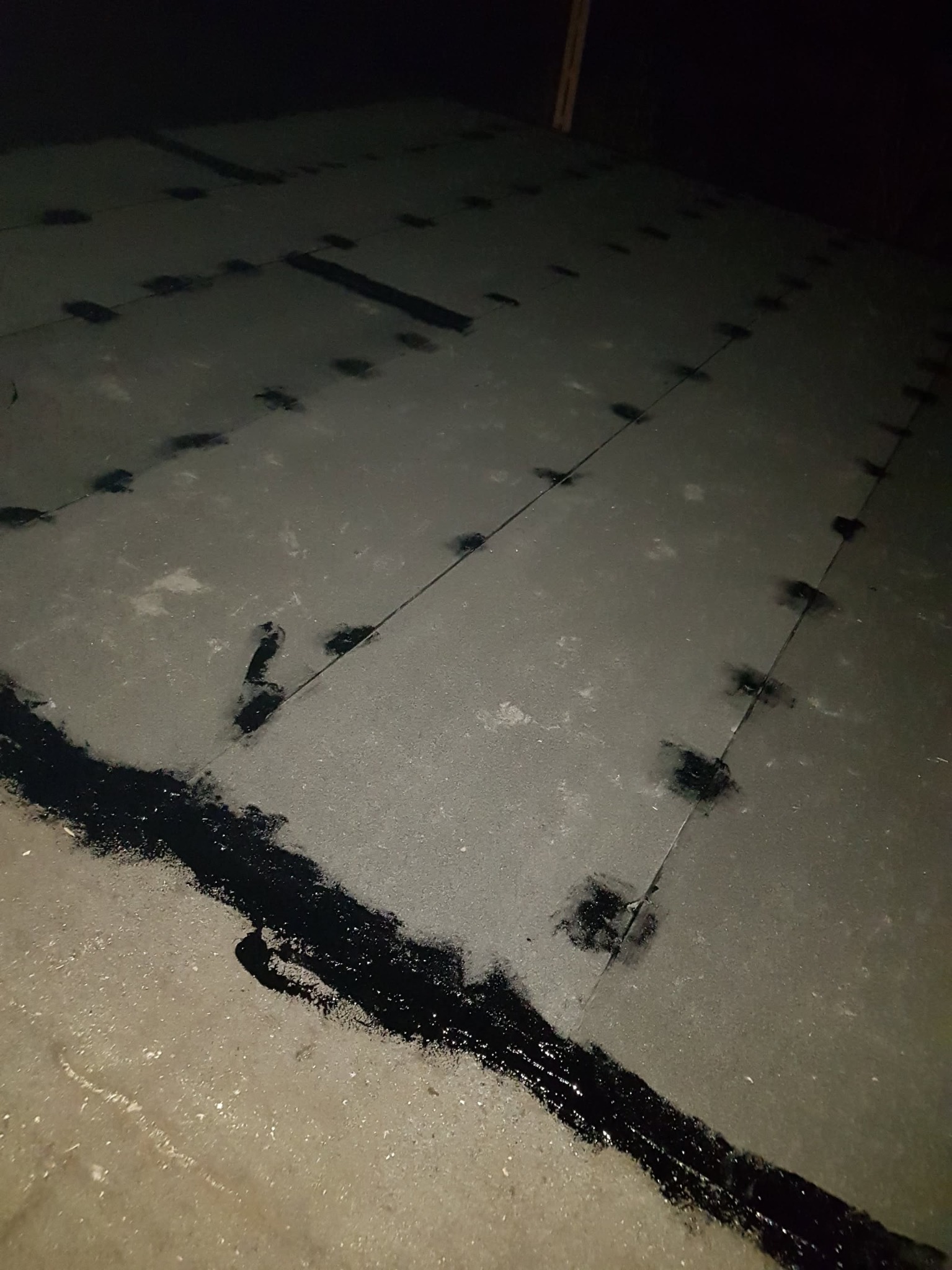 Szary betonowy podkład pod ogrzewanie podłogowe z czarnymi punktami i smugami uszczelniającymi, widoczne łączenia płyt.