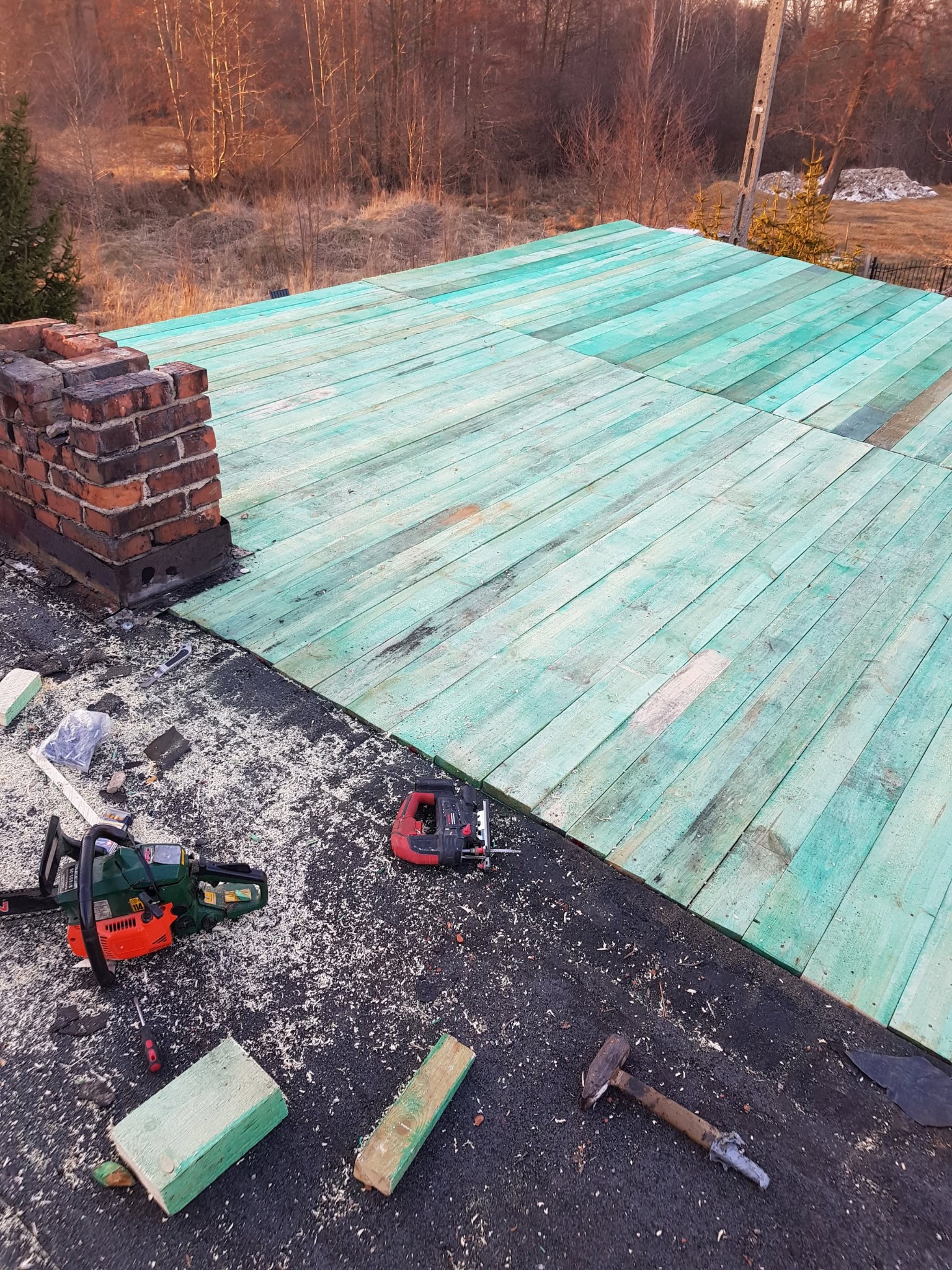 Fragment dachu pokryty zielonymi deskami, widoczny komin z czerwonej cegły, piła łańcuchowa, wyrzynarka, młotek i drobne elementy budowlane na czarnej papie, w tle drzewa i pole.