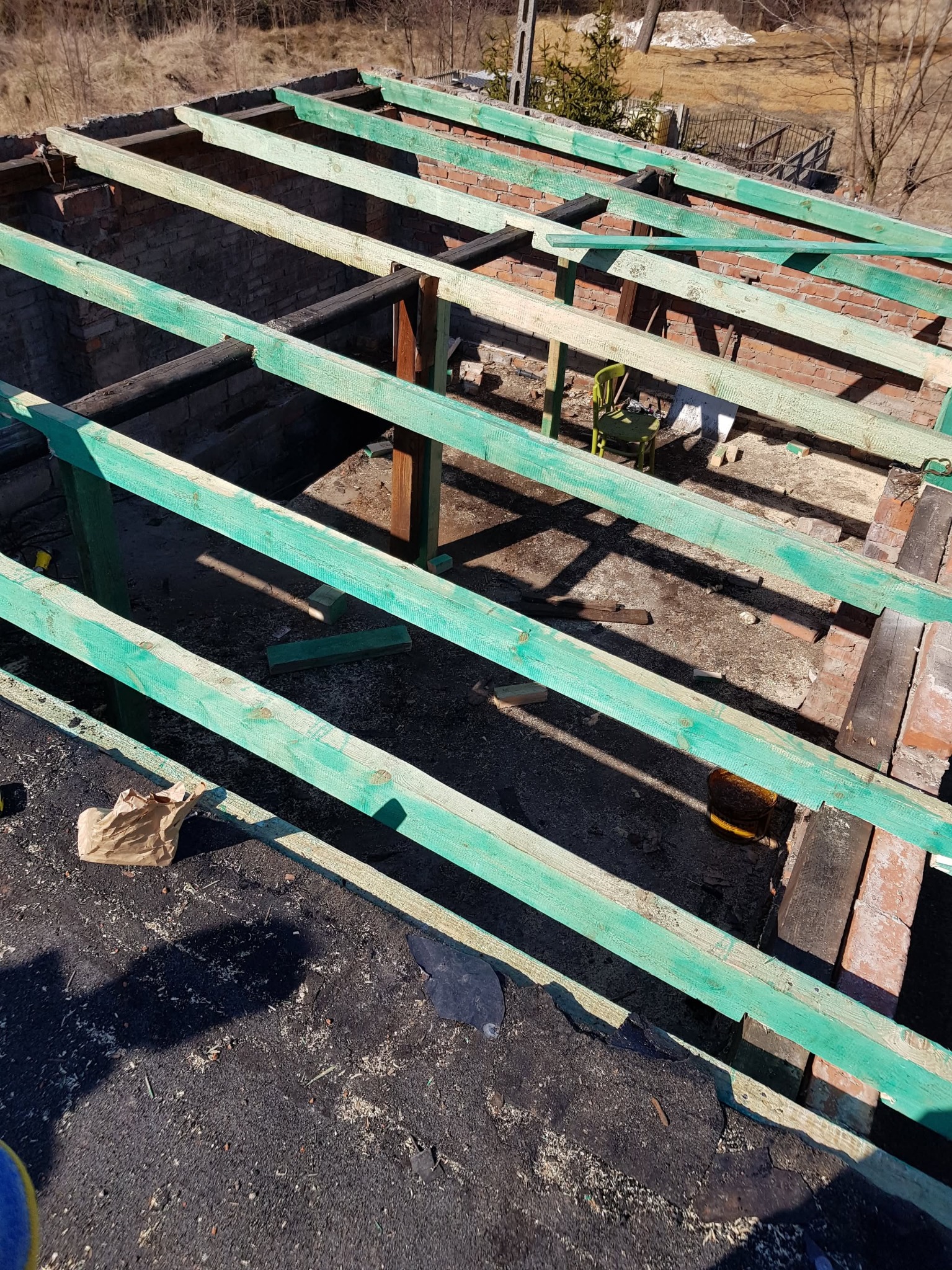 Konstrukcja więźby dachowej z pomalowanymi na zielono elementami drewnianymi, widoczna ceglana ściana budynku w tle, resztki materiałów budowlanych na dachu, perspektywa z góry.