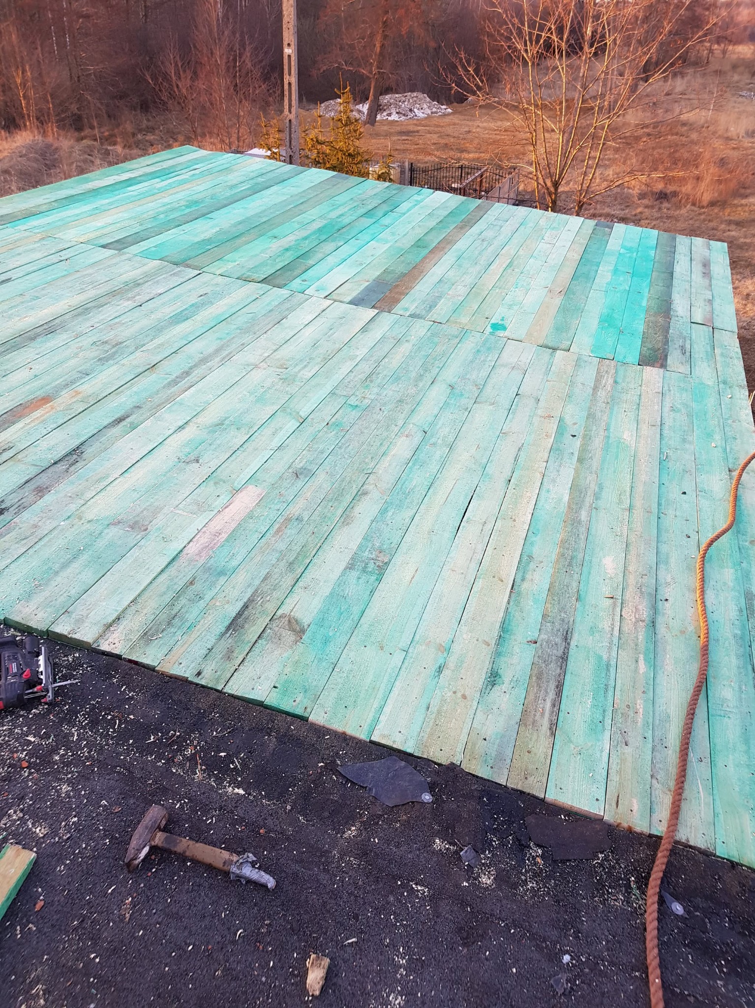 Widok z góry na drewnianą konstrukcję dachu pokrytą zieloną impregnacją, z widocznym młotkiem, narzędziami i liną na czarnym podłożu dachowym.