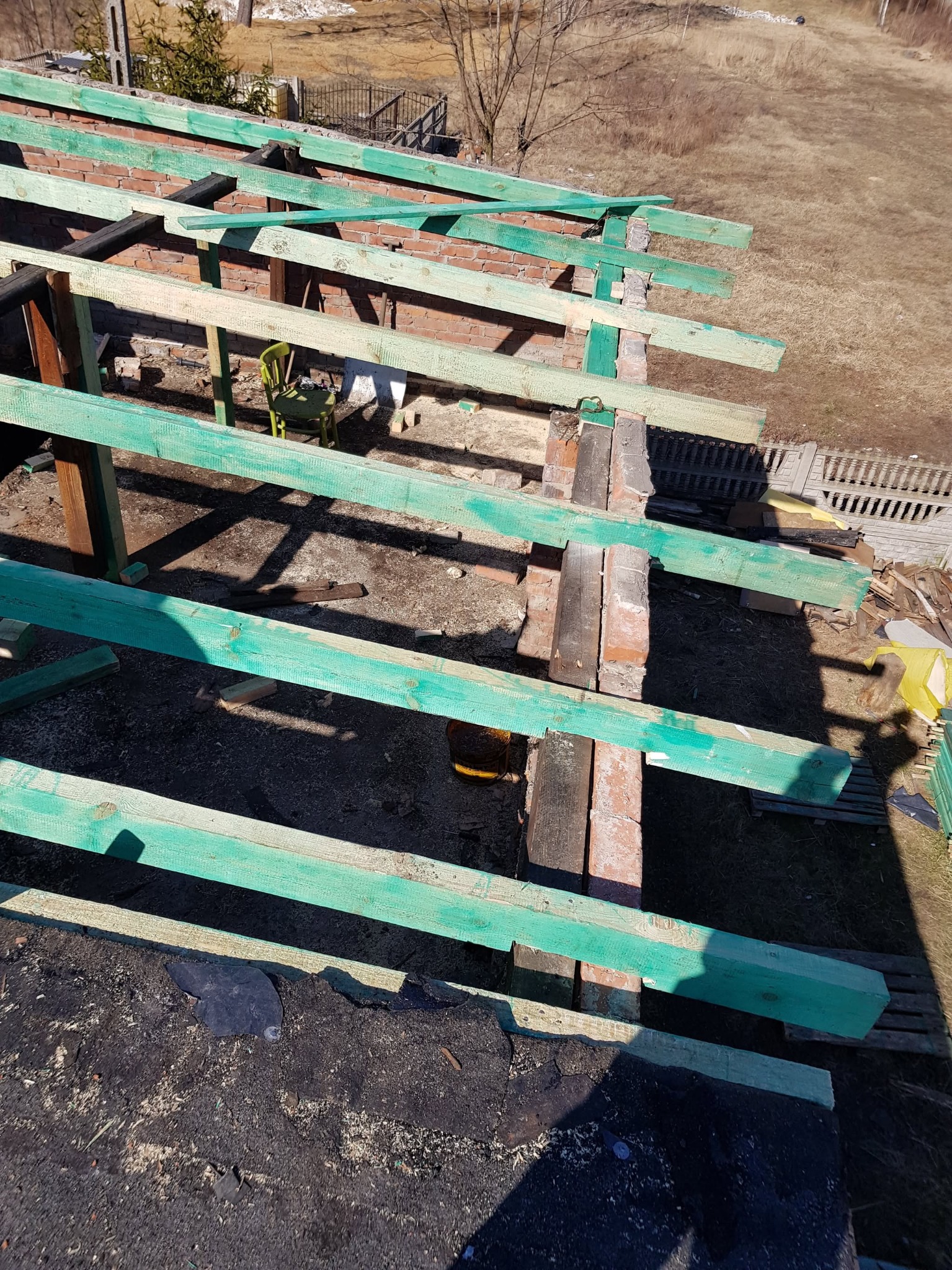 Konstrukcja dachu w trakcie budowy, z zielonymi belkami i ceglanymi kominami, widziana z góry, z fragmentem starego pokrycia dachowego w dolnej części kadru.