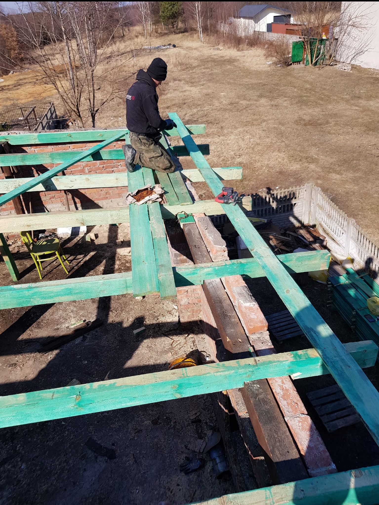 Budowa więźby dachowej z pomalowanymi na zielono elementami, widok z góry na dekarza w czapce i kurtce, pracującego na dachu z czerwonej cegły, z zielonym krzesłem i narzędziami w tle.