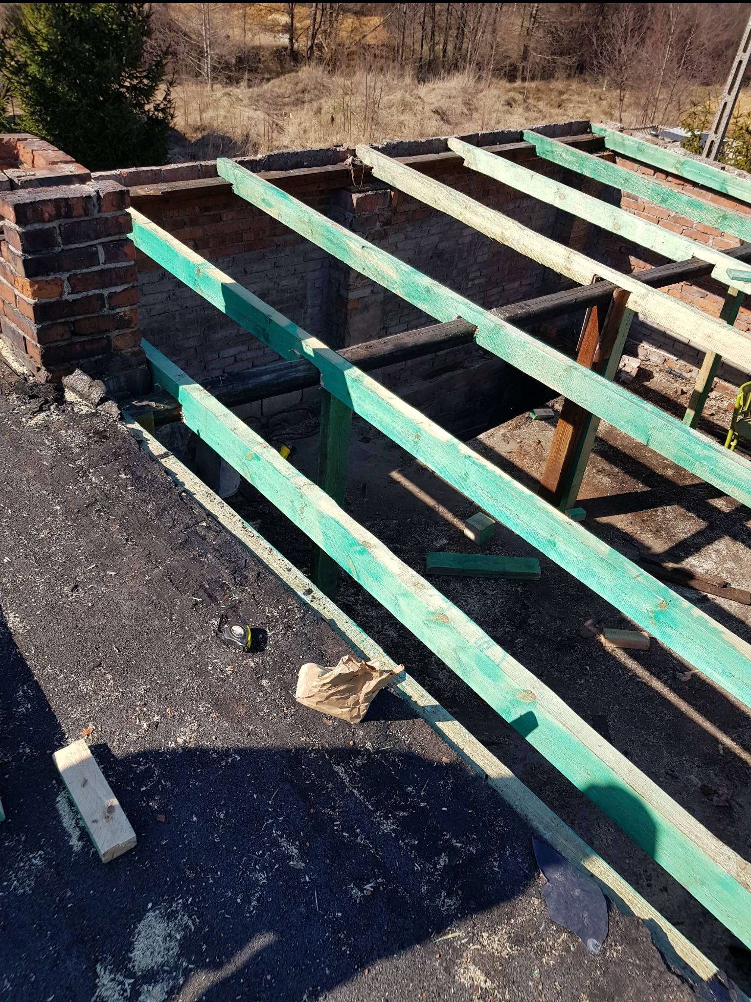 Konstrukcja dachu z pomalowanymi na zielono belkami, widoczny komin z cegły, narzędzia i resztki materiałów na czarnej papie dachowej.