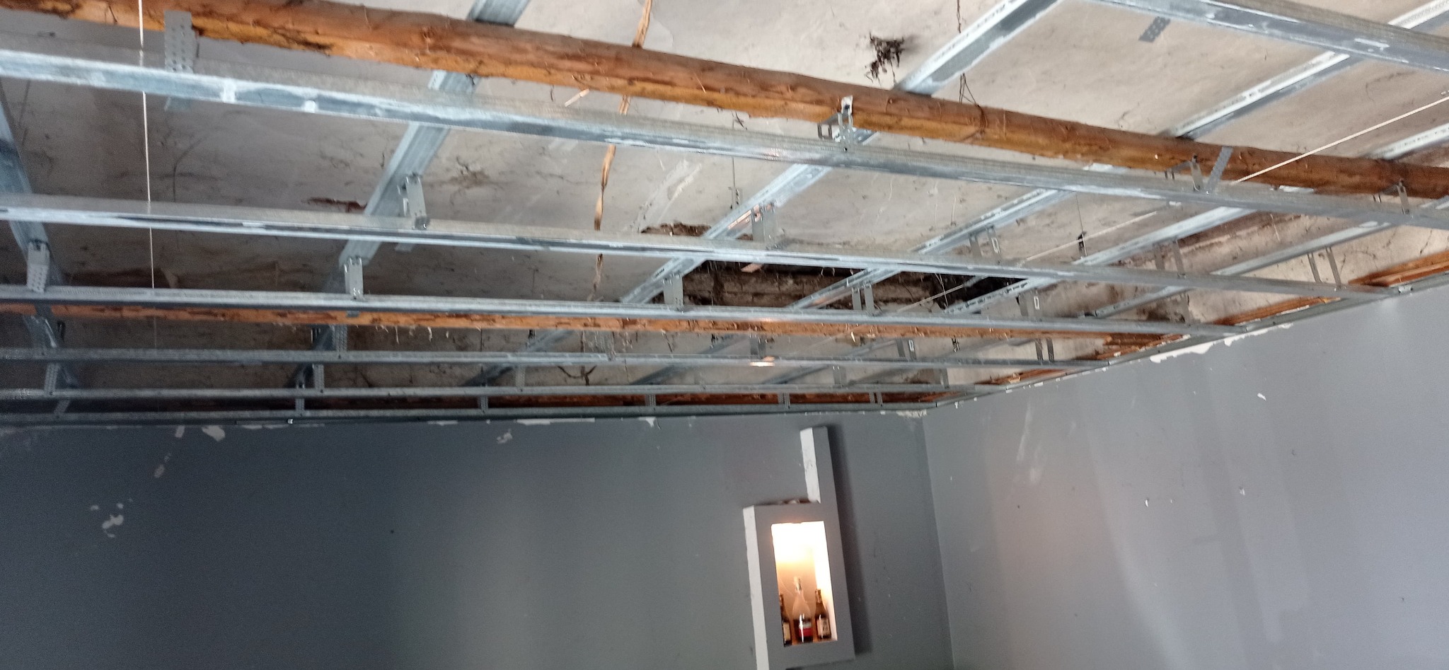 Stelaż sufitu podwieszanego z widocznymi starymi belkami stropowymi i fragmentem ściany pomalowanej na szaro z wnęką oświetleniową.