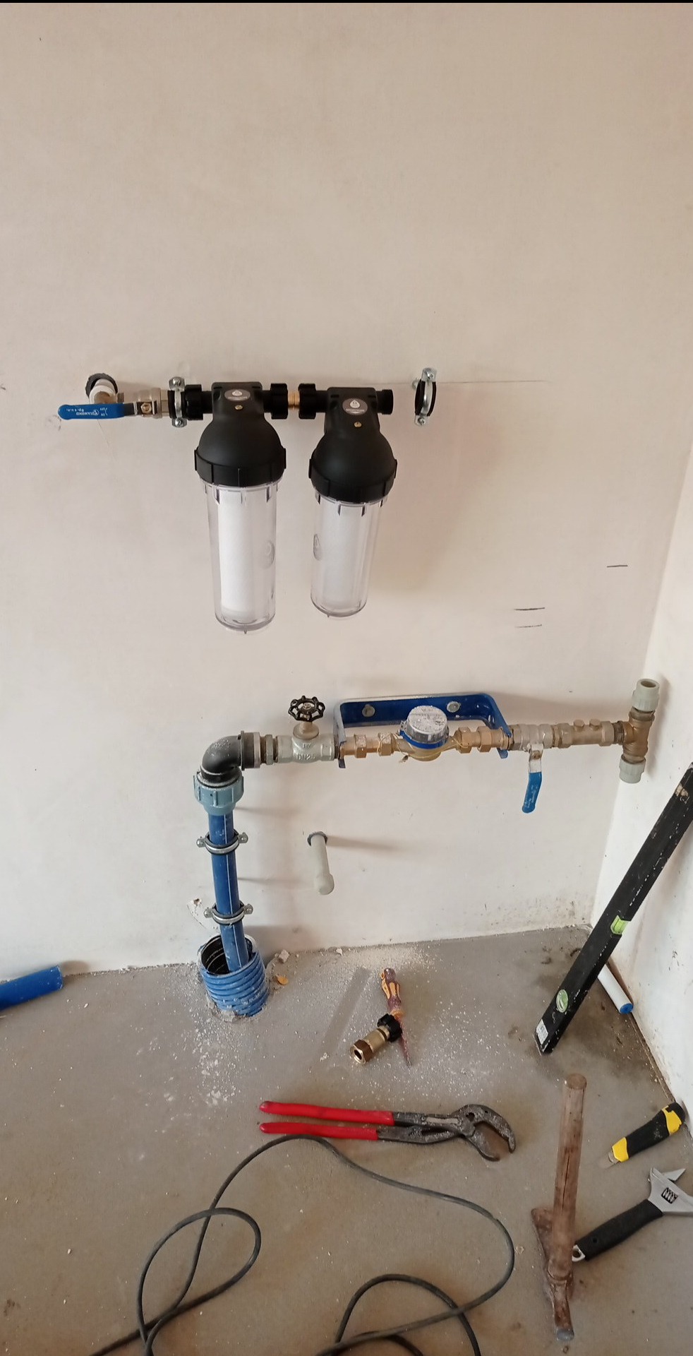 Montaż instalacji wodnej: widoczne dwa filtry wody z przezroczystymi obudowami, rury miedziane, licznik wody z niebieską obudową, zawory, oraz narzędzia hydrauliczne na podłodze z resztkami gruzu.