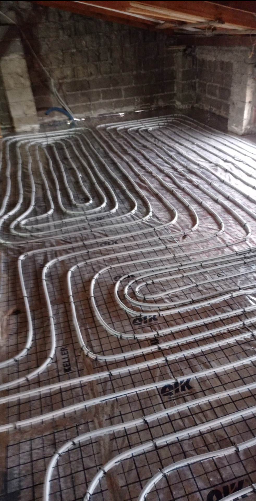 Instalacja ogrzewania podłogowego w surowym stanie, widoczne białe rury ułożone na czarnej siatce na brązowej folii, ściany z cegły, poddasze.