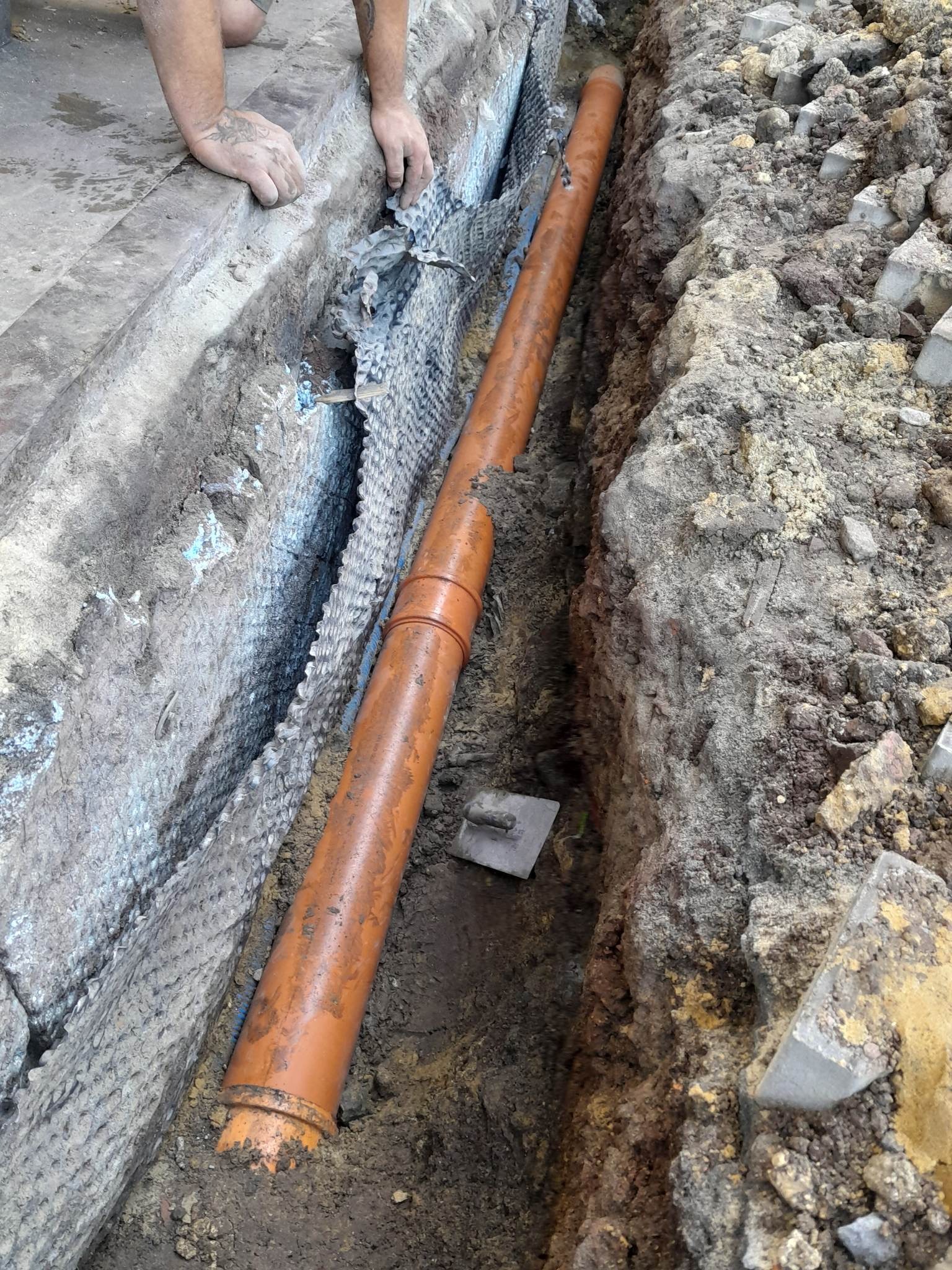 Pomarańczowa rura kanalizacyjna ułożona w wykopie ziemnym, zabezpieczonym geowłókniną; widoczne dłonie robotnika i kielnia.
