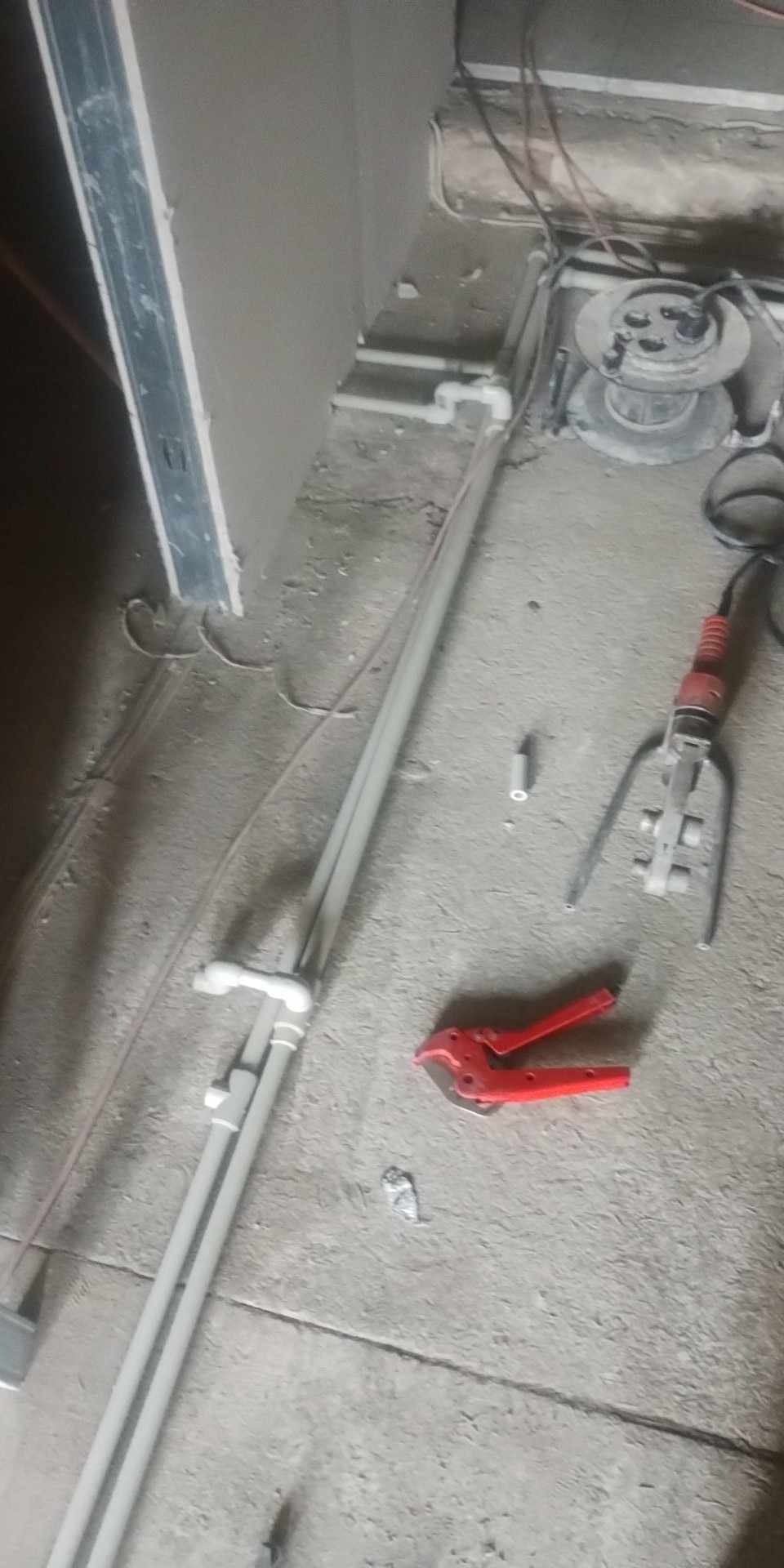 Przygotowanie instalacji hydraulicznej: białe rury z kształtkami, czerwone nożyce do cięcia rur, zgrzewarka do rur i fragment ściany w trakcie prac wykończeniowych.