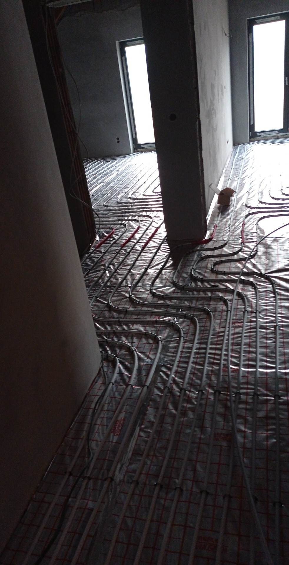 Montaż ogrzewania podłogowego: widoczne rury układane na folii z siatką w surowym wnętrzu z oknami.