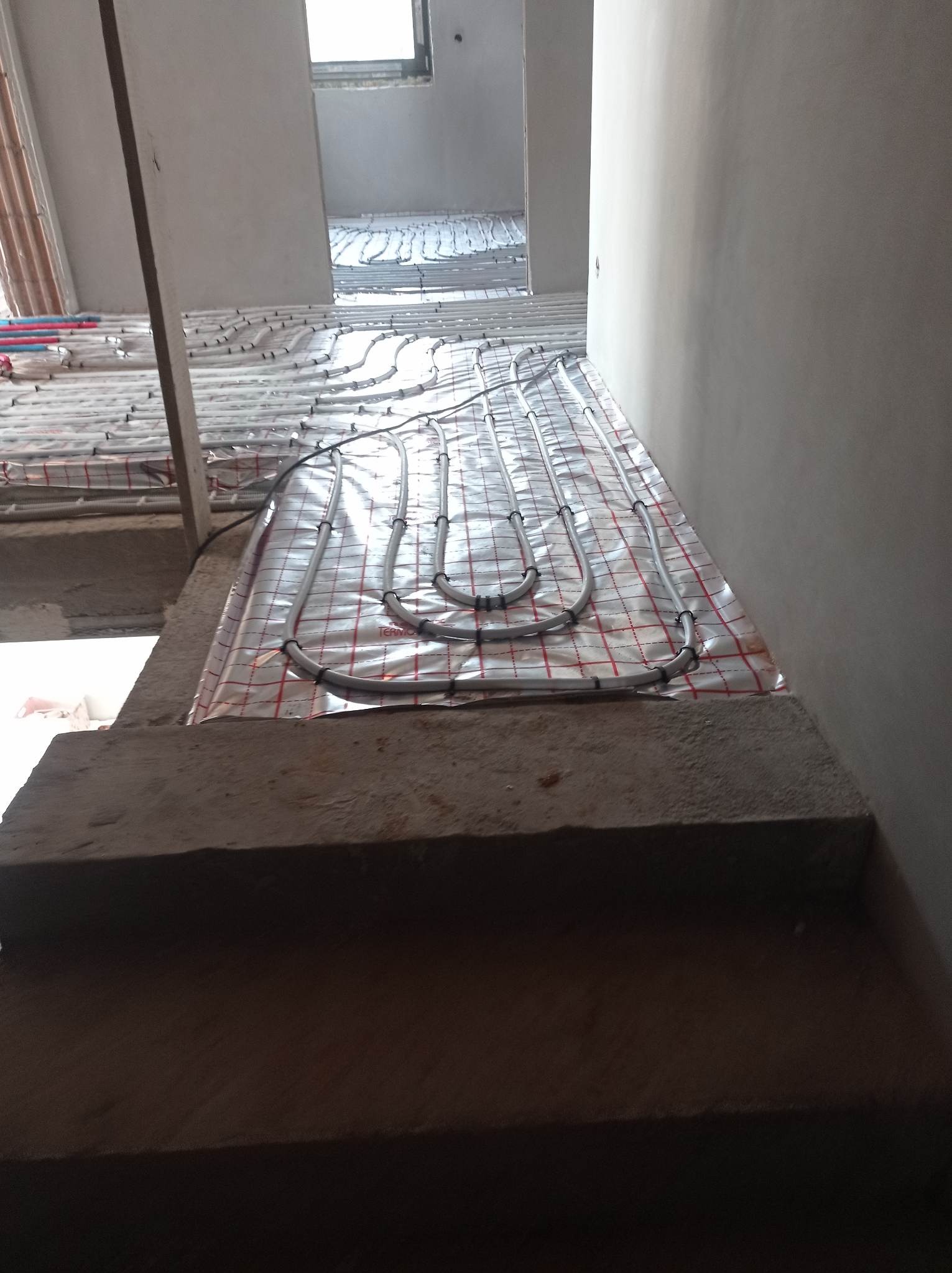Rury ogrzewania podłogowego ułożone na folii z siatką, widok z góry na betonowe schody w surowym wnętrzu.