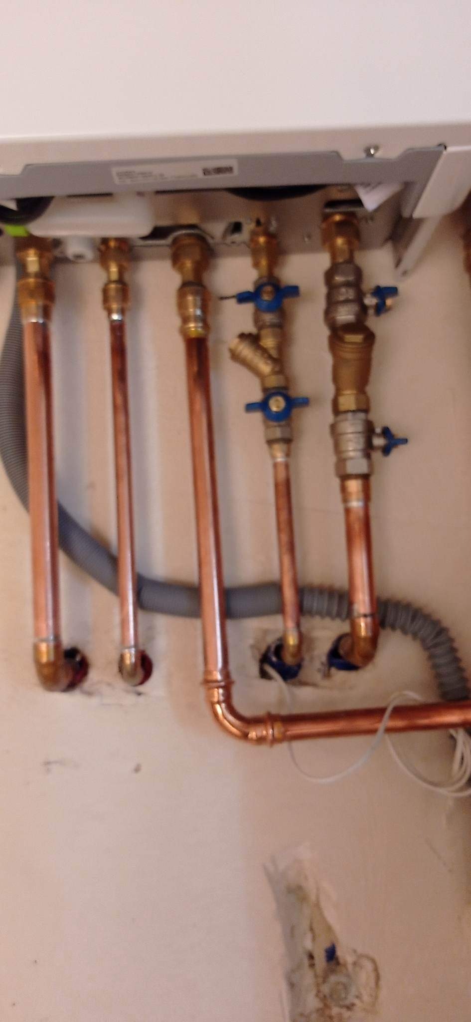 Miedziane rury instalacji grzewczej z zaworami, widoczne połączenia i fragment kotła, ściana z ubytkami po pracach instalacyjnych.