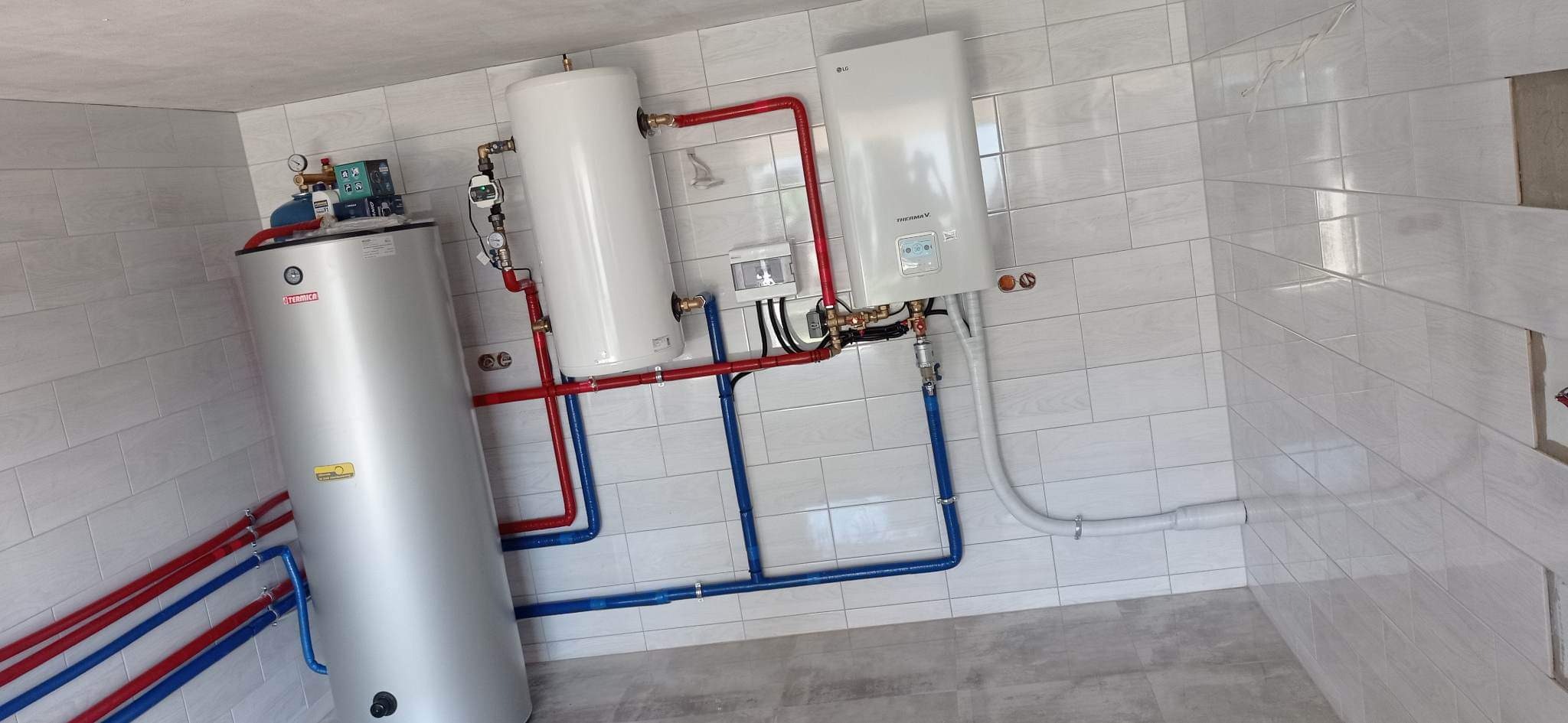 Zmontowana instalacja grzewcza z piecem gazowym, zasobnikiem ciepłej wody i bojlerem w pomieszczeniu wyłożonym białymi płytkami w Bytomiu, z widocznymi rurami w kolorach czerwonym i niebieskim.