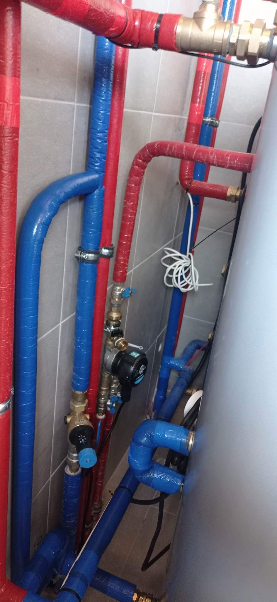 Instalacja grzewcza z czerwonymi i niebieskimi rurami zaizolowanymi termicznie, widoczne zawory, pompy obiegowe i zbiornik akumulacyjny na tle szarych płytek.