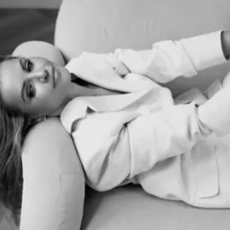 Czarno-białe, artystyczne ujęcie blond kobiety w białym garniturze, leżącej na jasnej sofie. Spojrzenie w obiektyw, subtelny uśmiech, miękkie światło.