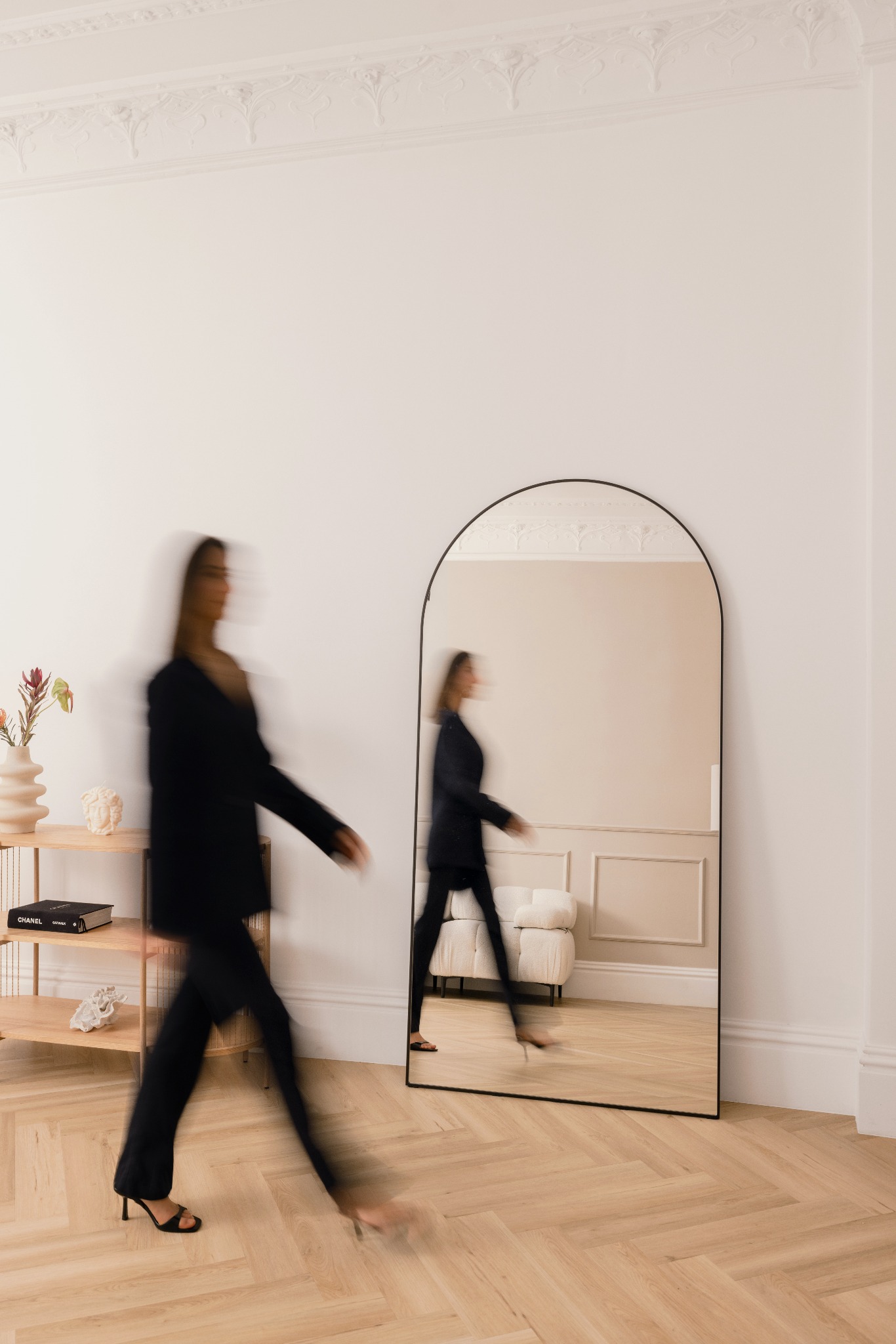 Rozmyta sylwetka kobiety w czarnym garniturze przechodzącej obok lustra łukowego, odbicie w lustrze, drewniana podłoga w jodełkę, półka z książkami i dekoracjami w tle, jasne wnętrze.