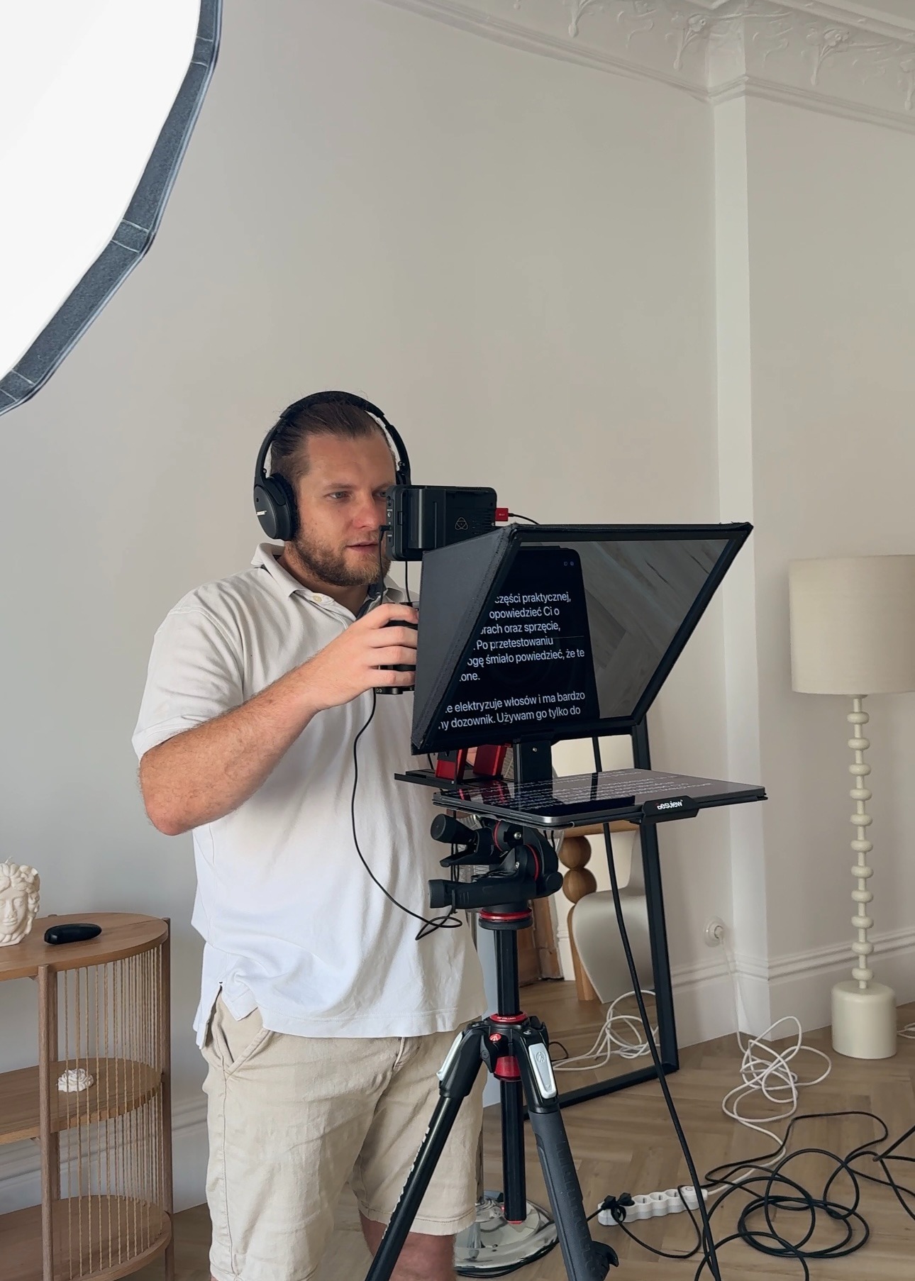 Mężczyzna w słuchawkach ustawia teleprompter z tekstem na ekranie, na statywie, w jasnym wnętrzu z oświetleniem studyjnym.