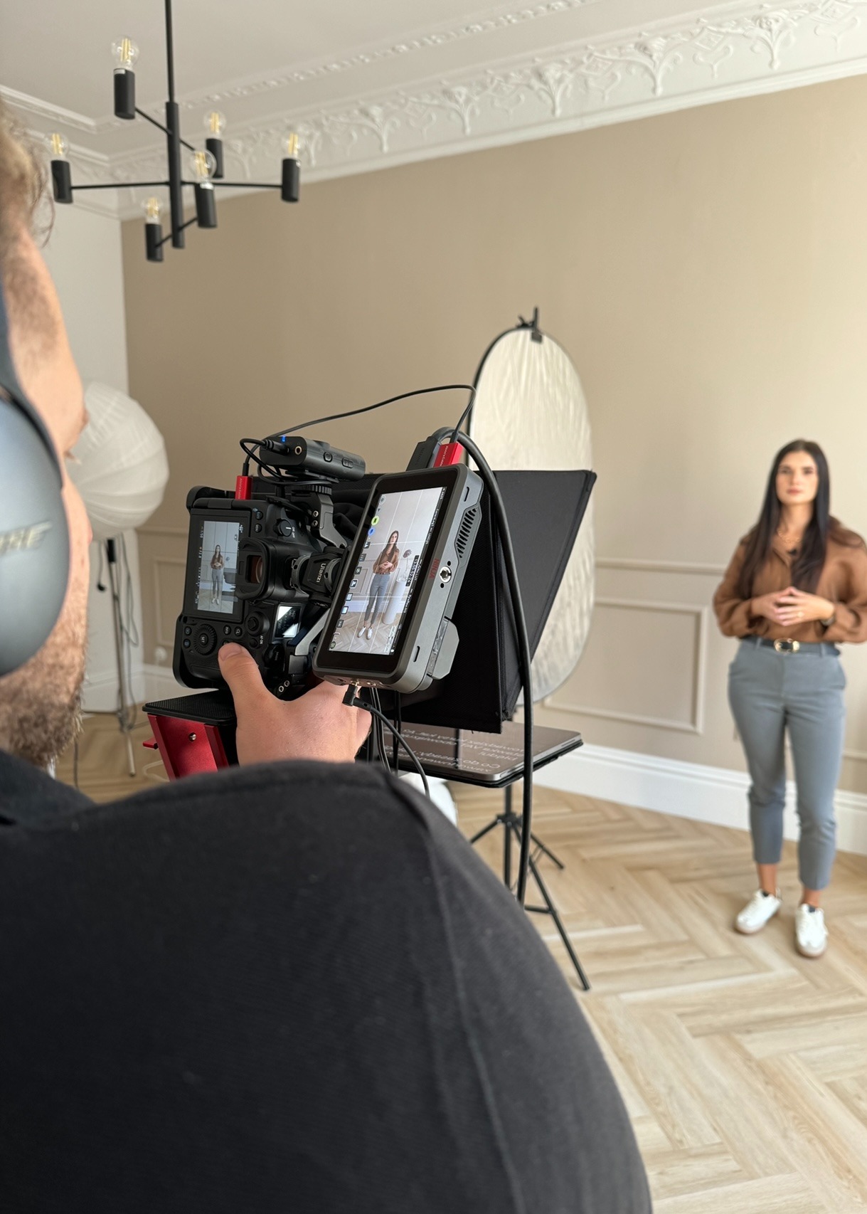 Kamera z podglądem kadrująca kobietę w koszuli i spodniach na tle jasnego pomieszczenia z lampą i blendą, widok zza operatora w słuchawkach.