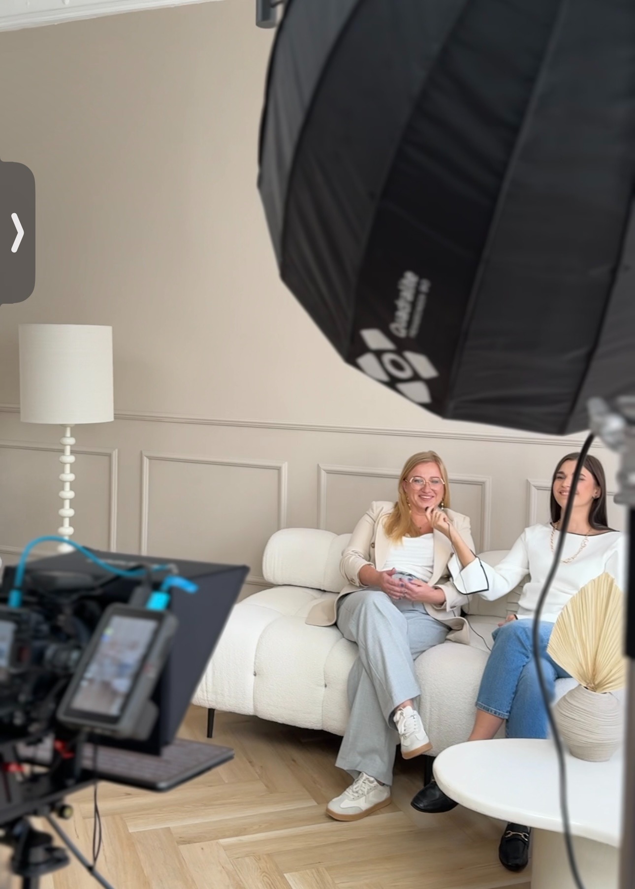Dwie kobiety siedzą na białej sofie podczas nagrywania wideo, widoczny profesjonalny sprzęt oświetleniowy i kamera na statywie.