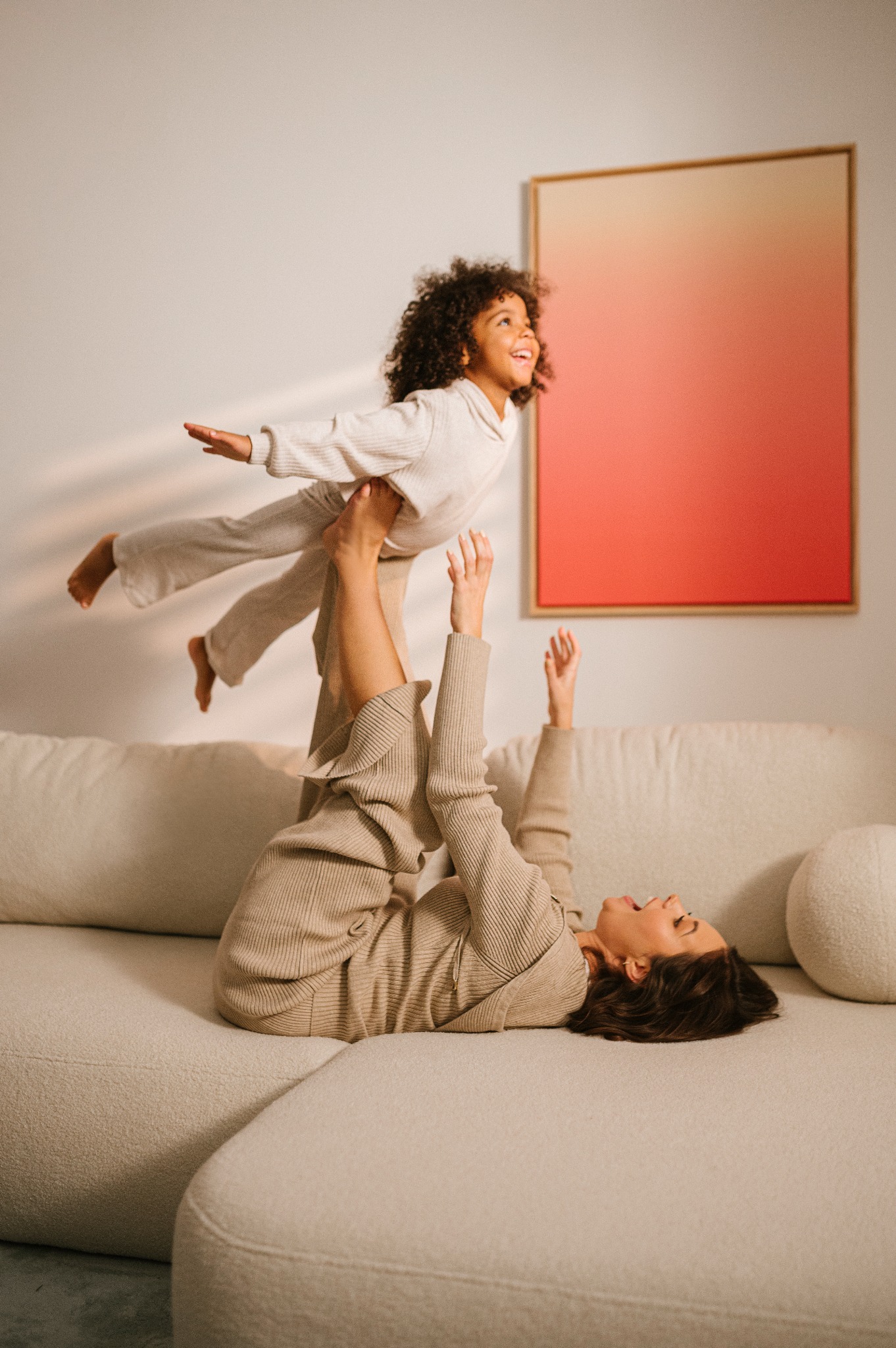 Matka leżąca na beżowej sofie, trzymająca w górze uśmiechniętą córkę z kręconymi włosami, na ścianie obraz z gradientem kolorów.