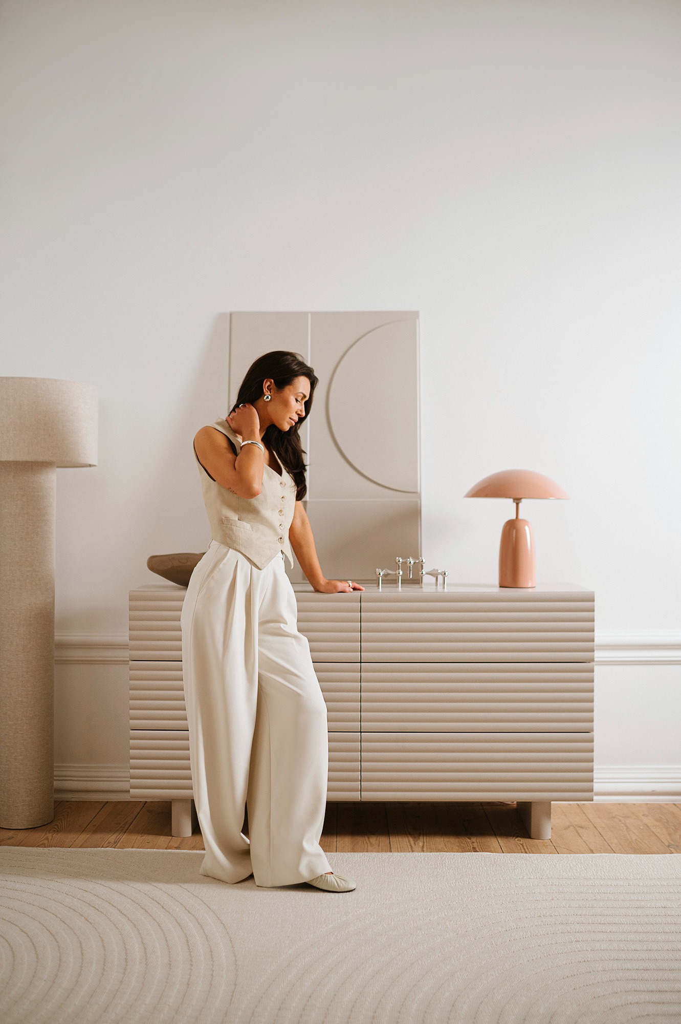 Kobieta w eleganckim stroju pozuje obok beżowej komody z lampą w minimalistycznym wnętrzu, podkreślając harmonię kolorów i faktur.