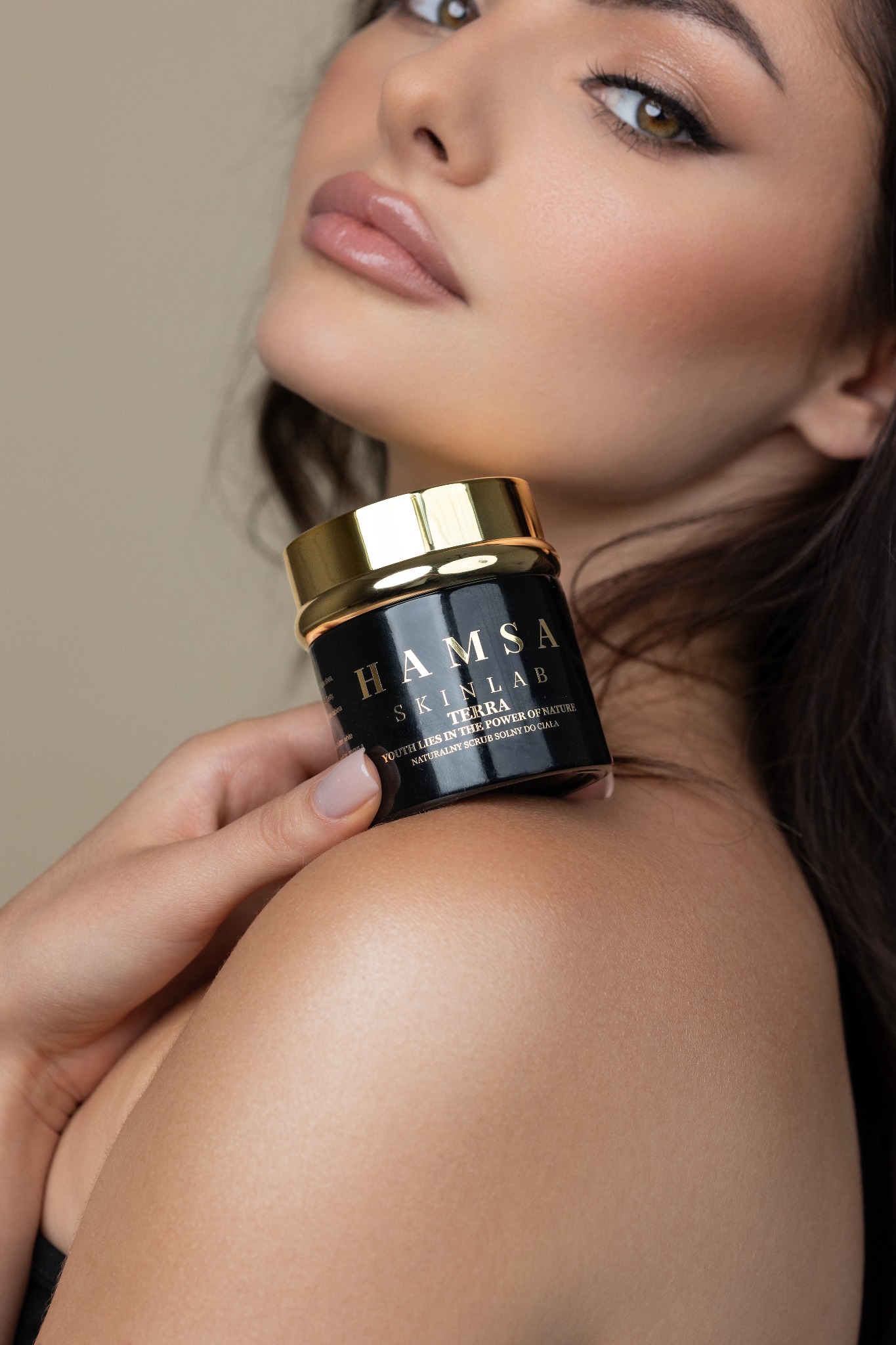 Kobieta z makijażem prezentuje czarny słoiczek z napisem HAMSA SKINLAB TERRA, naturalny scrub solny do ciała, trzymając go na ramieniu.