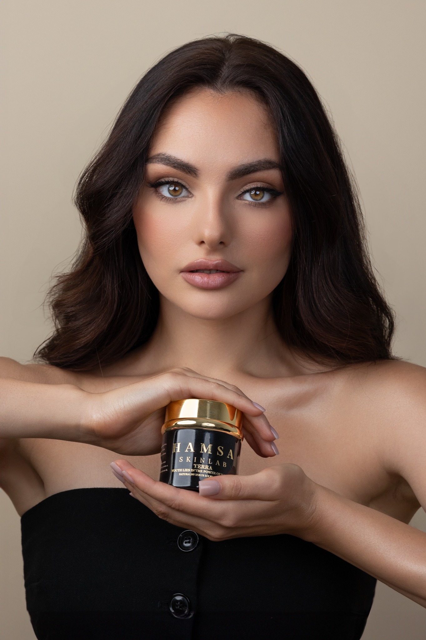 Portret kobiety trzymającej słoiczek z kremem do twarzy marki Hamsa Skinlab na dłoniach, z delikatnym makijażem i długimi, ciemnymi włosami na beżowym tle.