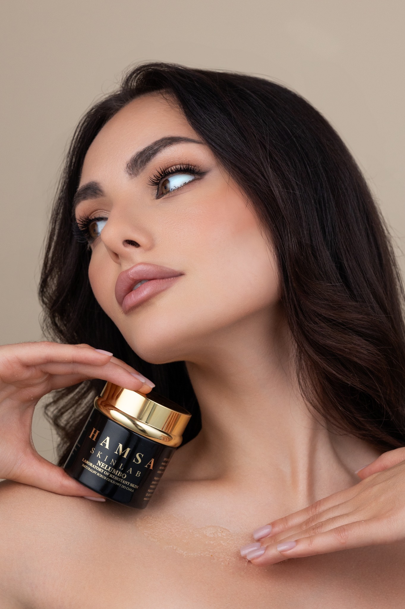 Kobieta z ciemnymi włosami trzyma czarny słoiczek z kremem marki Hamsa Skinlab Nelumbo B i aplikuje go na dekolt, spoglądając w górę w lewo.