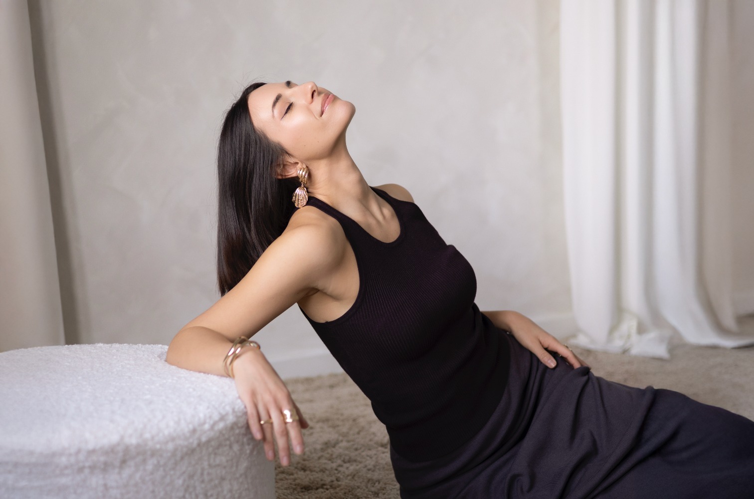 Portret kobiety w czarnym topie i długiej spódnicy, z biżuterią w stylu vintage, siedzącej na podłodze z zamkniętymi oczami, w naturalnym oświetleniu.