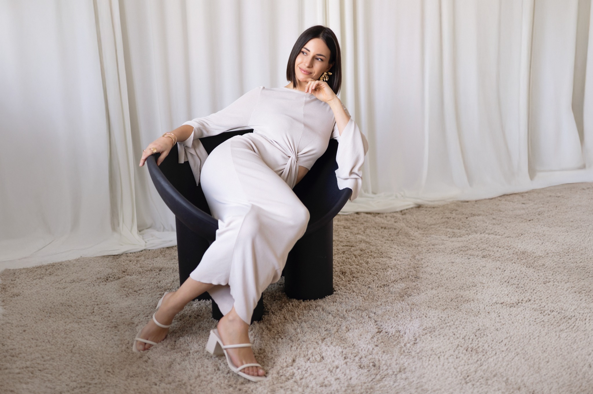 Elegancka kobieta w białym kombinezonie pozuje siedząc w czarnym fotelu na tle białych zasłon i beżowego dywanu, patrząc w bok z uśmiechem.