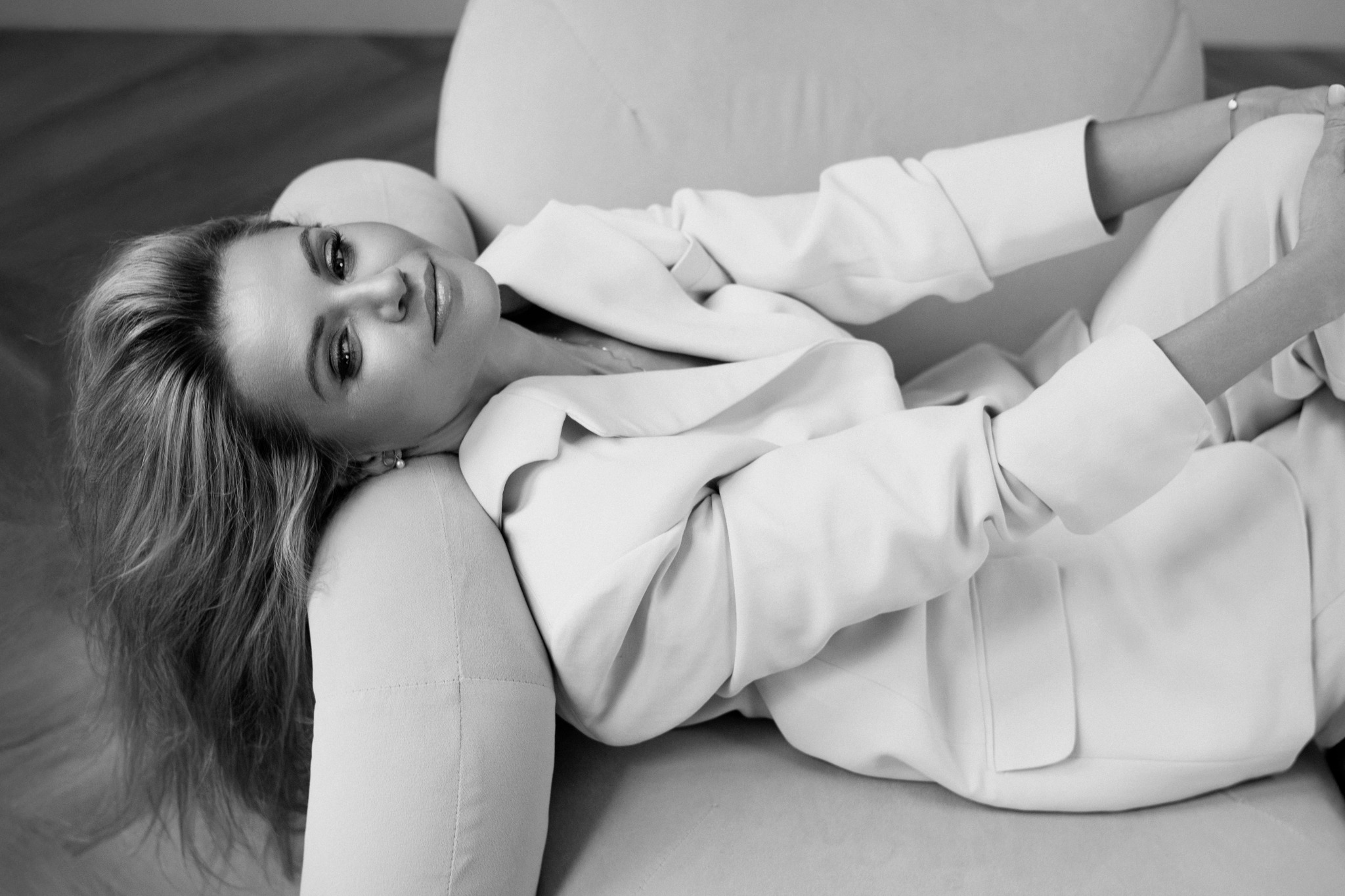 Czarno-białe, artystyczne ujęcie blond kobiety w białym garniturze, leżącej na jasnej sofie. Spojrzenie w obiektyw, subtelny uśmiech, miękkie światło.
