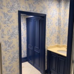 LW DECOR - ŁUKASZ WYMYSŁOWSKI - Elegancka łazienka z granatowymi drzwiami i szafką umywalkową, ściany pokryte tapetą w stylu toile de jouy, beżowa umywalka i płytki podłogowe.