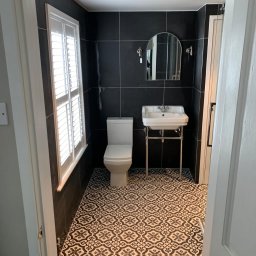 LW DECOR - ŁUKASZ WYMYSŁOWSKI - Nowoczesna łazienka z czarnymi płytkami ściennymi, wzorzystą czarno-białą podłogą, białą toaletą i umywalką na chromowanym stelażu, oświetlona przez okno z żaluzjami.