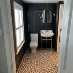 Nowoczesna łazienka z czarnymi płytkami ściennymi, wzorzystą czarno-białą podłogą, białą toaletą i umywalką na chromowanym stelażu, oświetlona przez okno z żaluzjami.