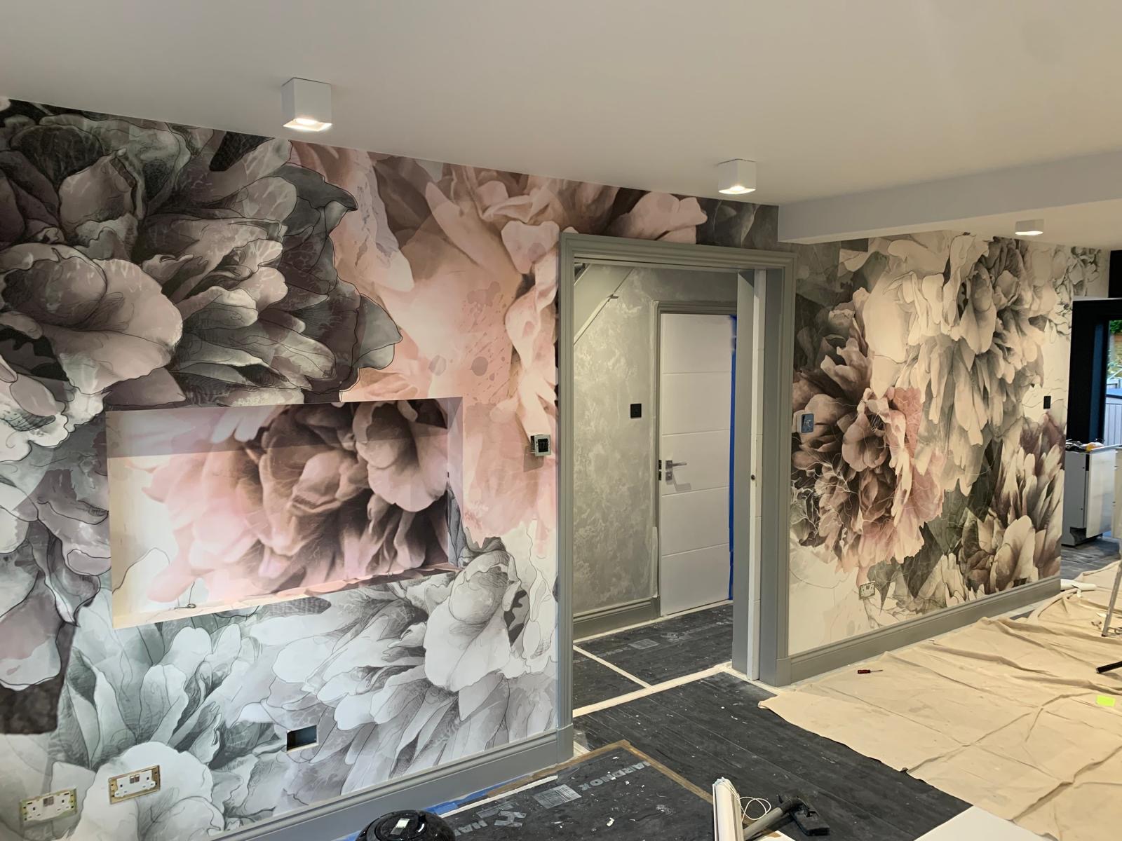 Pokój po remoncie z fototapetą w motywy kwiatowe w odcieniach szarości i różu, widoczna wnęka na telewizor, szara stolarka drzwiowa i zabezpieczona podłoga.