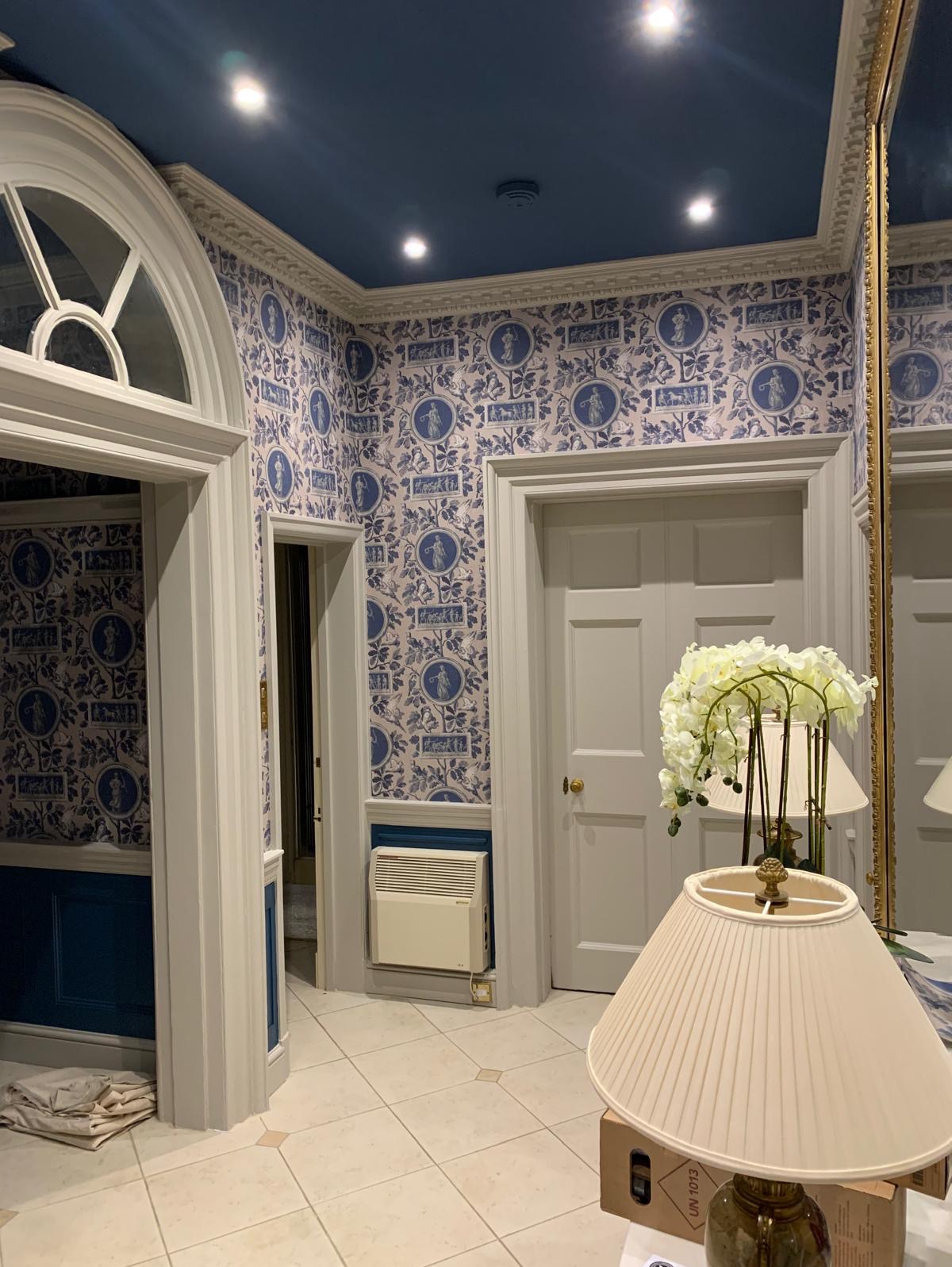 Wnętrze przedpokoju z wzorzystą tapetą w odcieniach bieli i niebieskiego, białe drzwi, grzejnik elektryczny na ścianie i lampa stołowa z kremowym abażurem na pierwszym planie.