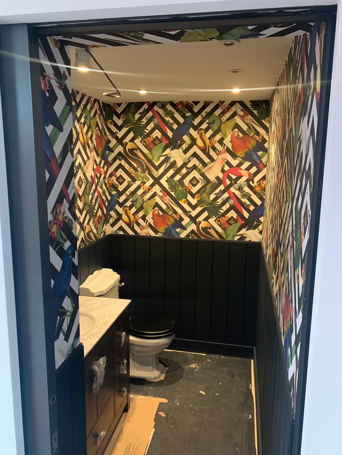 Ukończona aranżacja małej łazienki z czarną boazerią i tapetą w tropikalne ptaki na białym tle w geometryczne wzory, widoczna toaleta i umywalka z szafką w stylu retro.